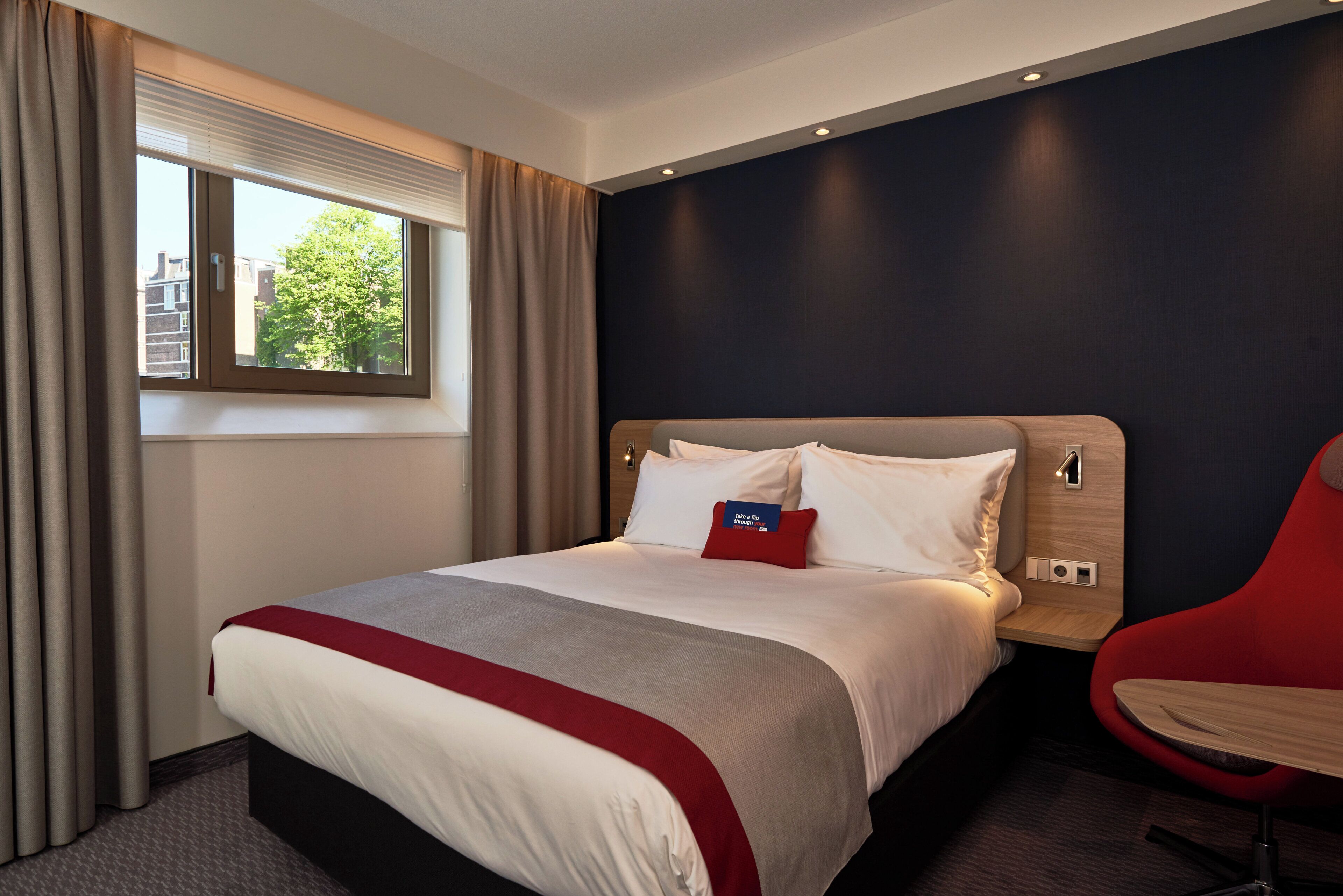 A hotel room with a bed, a window with curtains, a red chair, and a wall-mounted light.