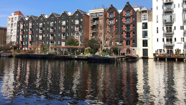 A row of multi-story buildings with balconies, a canal in the foreground, and a clear blue sky. - Holiday Inn Express Amsterdam - City Hall by IHG (Amsterdam)
