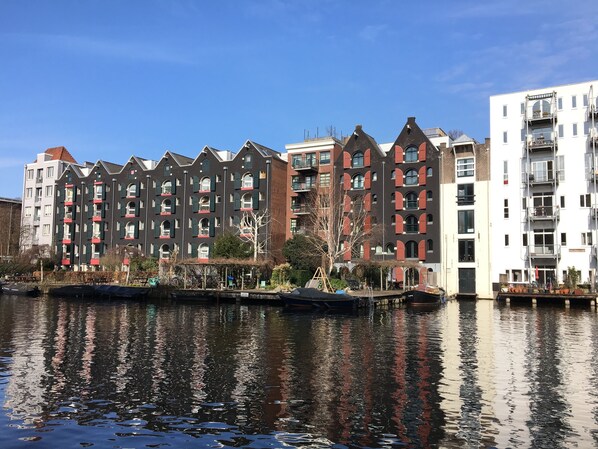 A row of multi-story buildings with balconies, a canal in the foreground, and a clear blue sky.
