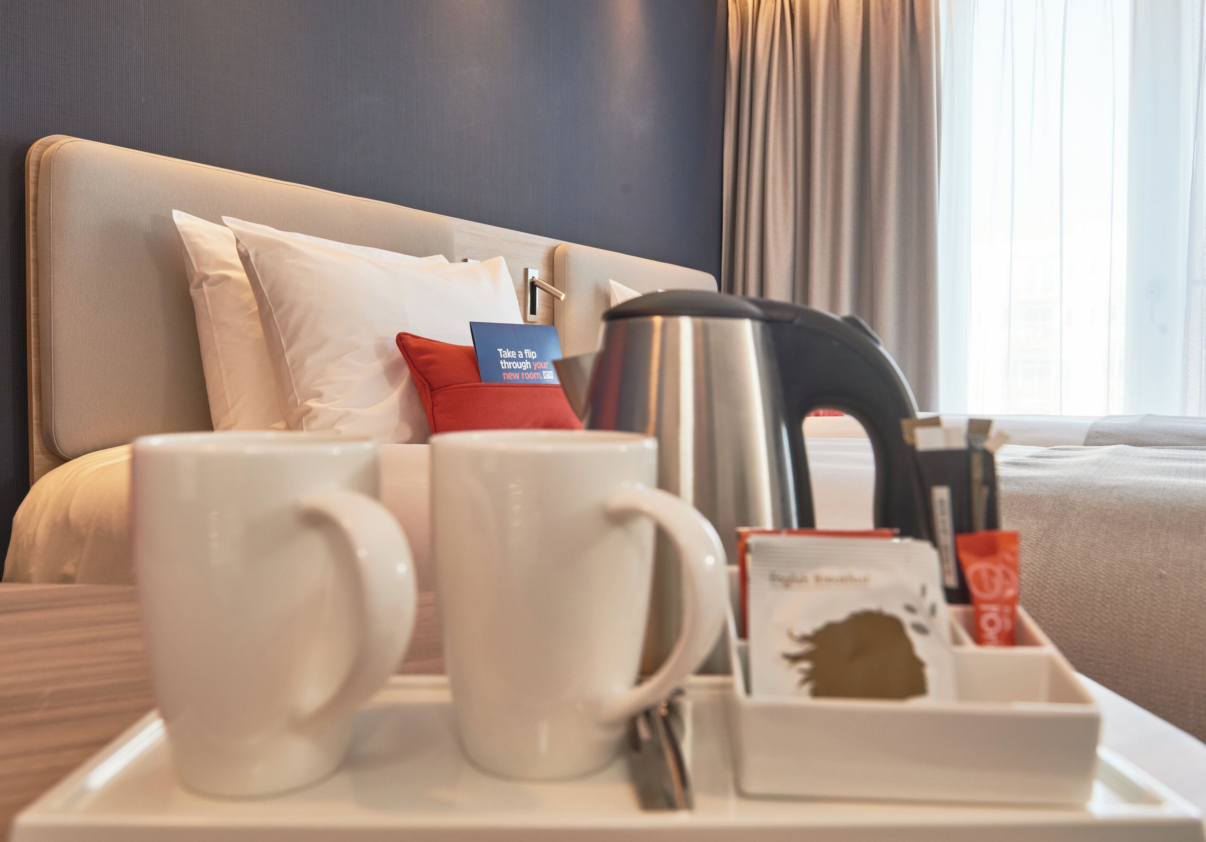 Habitació d'hotel ben ordenada amb un llit, almoines, una safata amb dues tasses blanques, una cafetera i una altra safata petita amb un paquet de te i un objecte vermell.