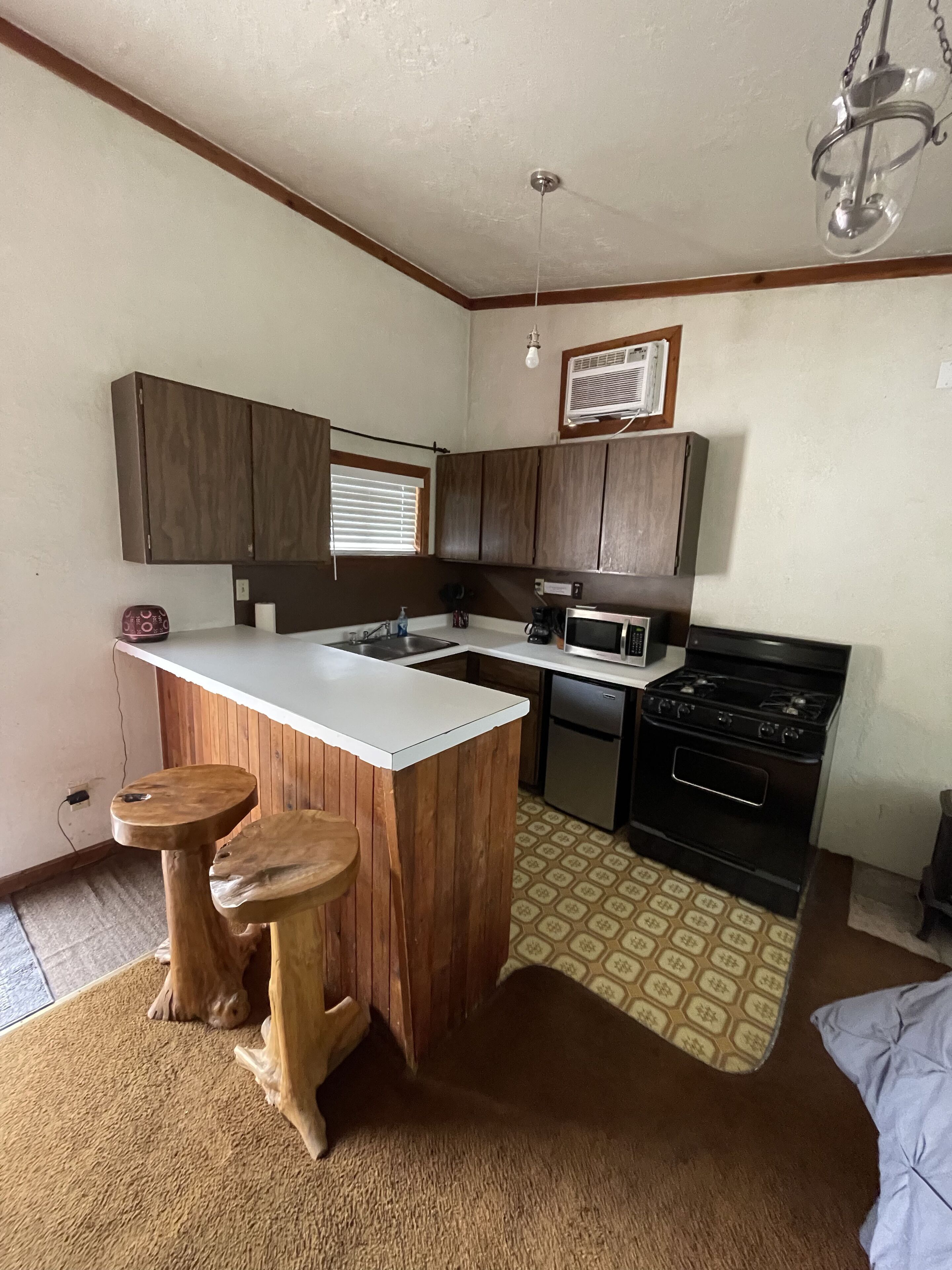 A compact kitchen with wooden cabinets, a white countertop, a built-in stove, and two stools.