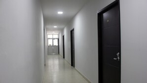 Un pasillo largo y estrecho con paredes blancas y piso de baldosas. Hay varias puertas a lo largo del pasillo, incluyendo una puerta negra con el número 106. El pasillo conduce a una puerta al final.