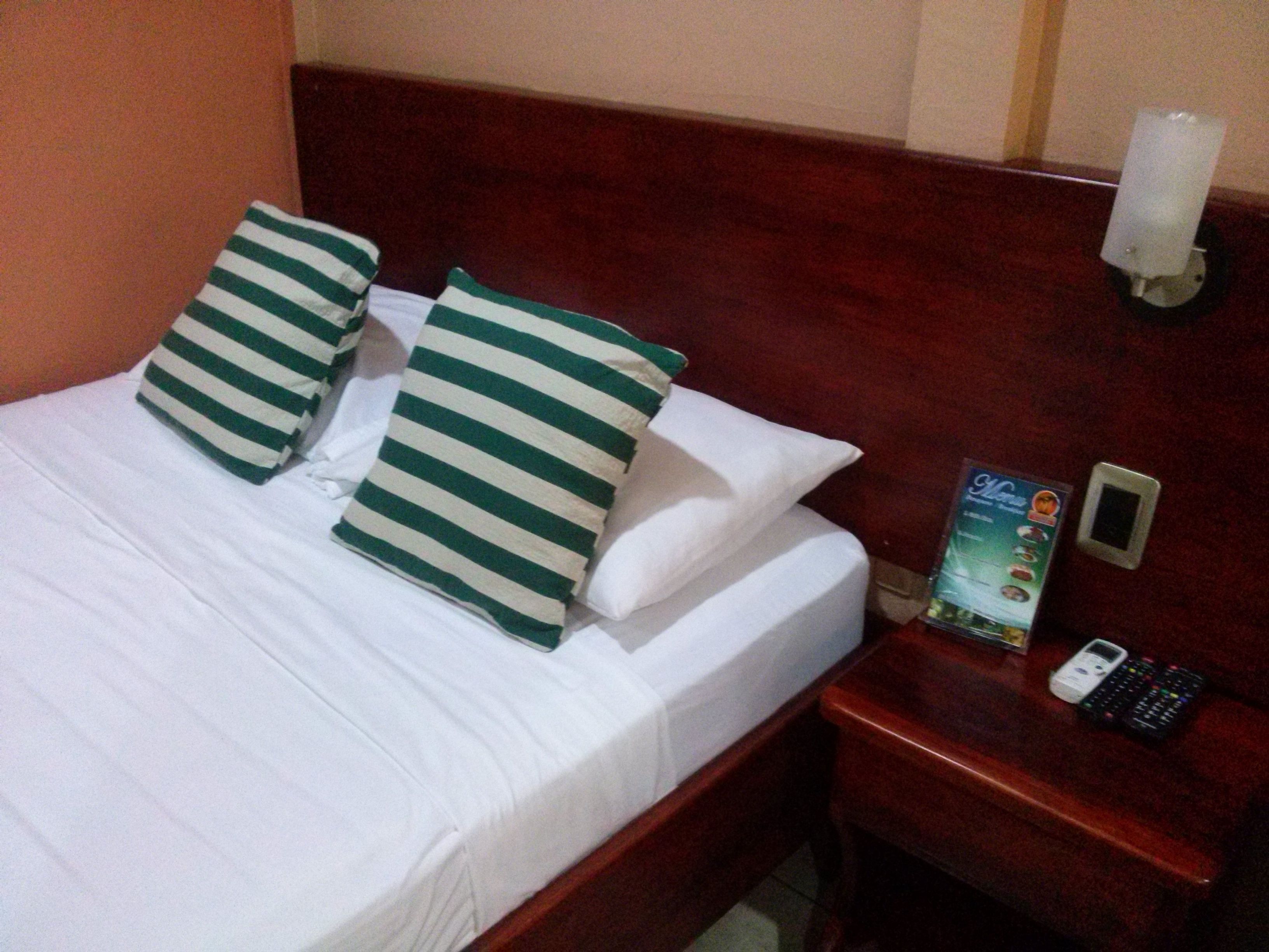 A hotel room with a neatly made bed, striped pillows, a wooden headboard, a nightstand with a remote control, and a brochure.
