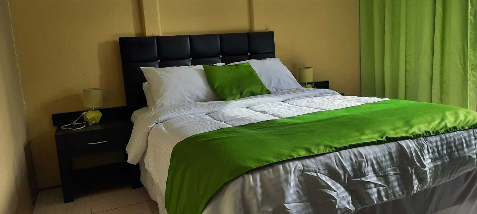 Una cama con cabecera negra, ropa de cama blanca y verde, y una cortina verde a la derecha.