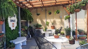 Un patio exterior con una pérgola, plantas en macetas, una mesa blanca y sillas negras.