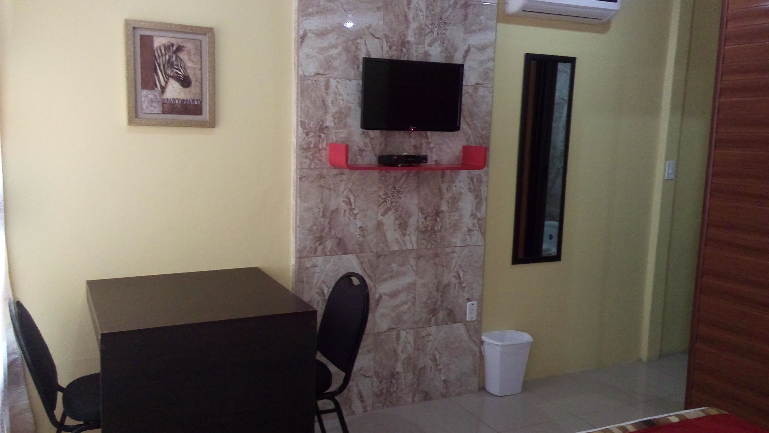 A small room with a table and chairs, a mounted TV, a trash bin, and a framed picture on the wall.