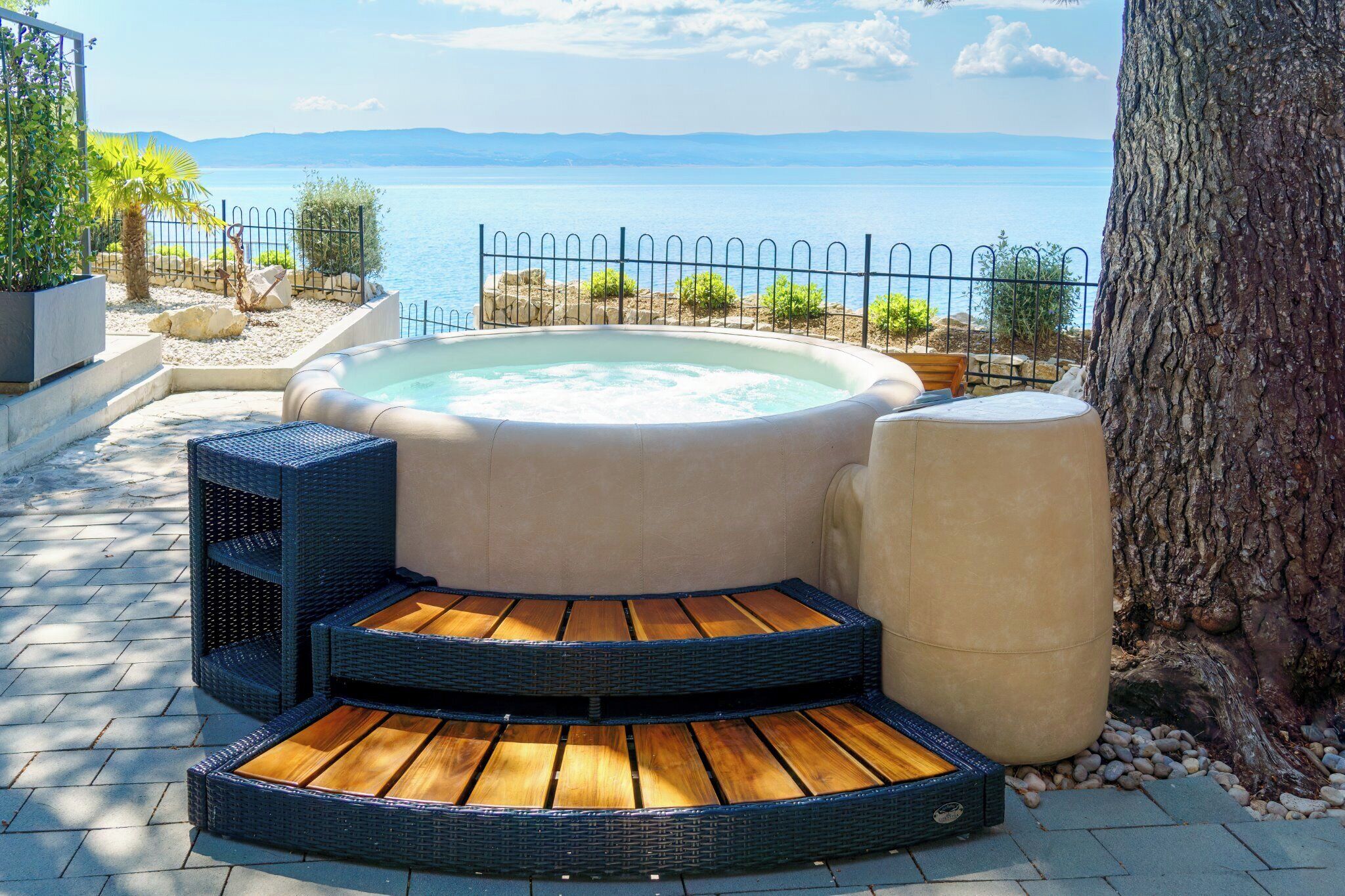 A hot tub with a wooden deck, surrounded by a fence, with a view of the sea and mountains in the background.