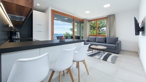 A modern kitchen with a built-in stove, white chairs, a gray sofa, and a view of the outdoors. - Apartments Bikin (Brela)