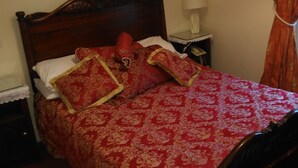 Una cama con colcha y almohadas rojas estampadas, cabecera de madera, mesita de noche con lámpara y espejo en la pared.