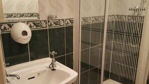 Un baño con lavamanos blanco, dispensador de papel higiénico con carita sonriente y zona de ducha con rejilla metálica.