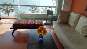 Una sala moderna con un sofá, un reposapiés estampado y una mesita con un jarrón de flores. Un ventanal grande ofrece vista al océano y palmeras.