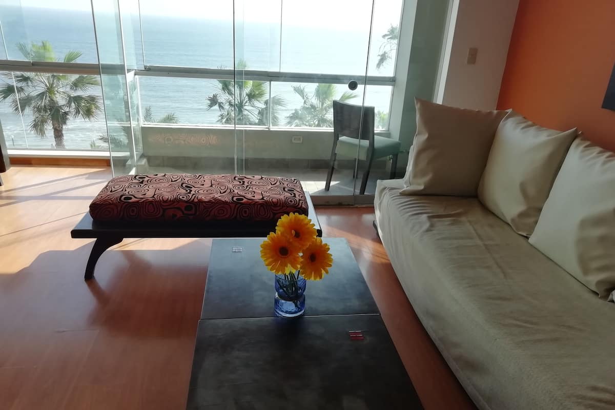 A modern living room with a sofa, a patterned ottoman, and a small table with a vase of flowers. A large window offers a view of the ocean and palm trees.