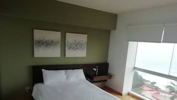 A bedroom with a large bed, two wall-mounted abstract paintings, a bedside table with a lamp, and a view of a park through a large window.