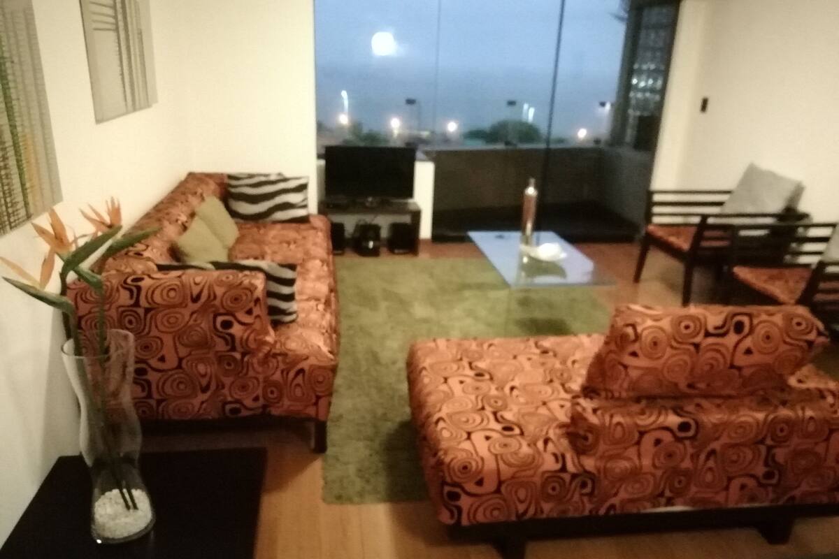 A living room with patterned sofas, a glass coffee table, and a view of the city at dusk.
