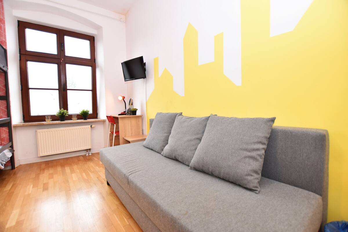 Une pièce avec un canapé gris, une fenêtre avec des stores, un téléviseur à écran plat fixé au mur et un mur jaune orné d’une fresque représentant un paysage urbain.