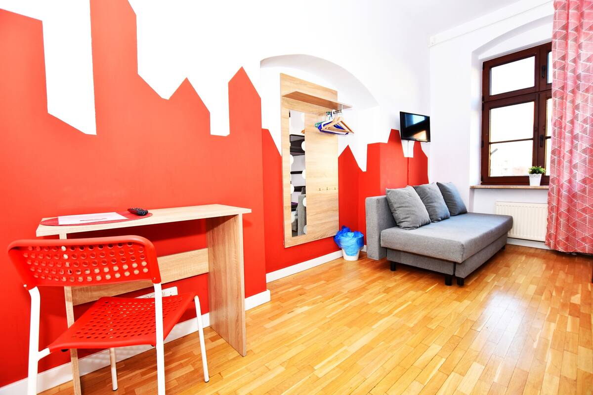 Une pièce avec un bureau en bois, une chaise rouge, un canapé gris et une fenêtre ornée d’un rideau rouge.