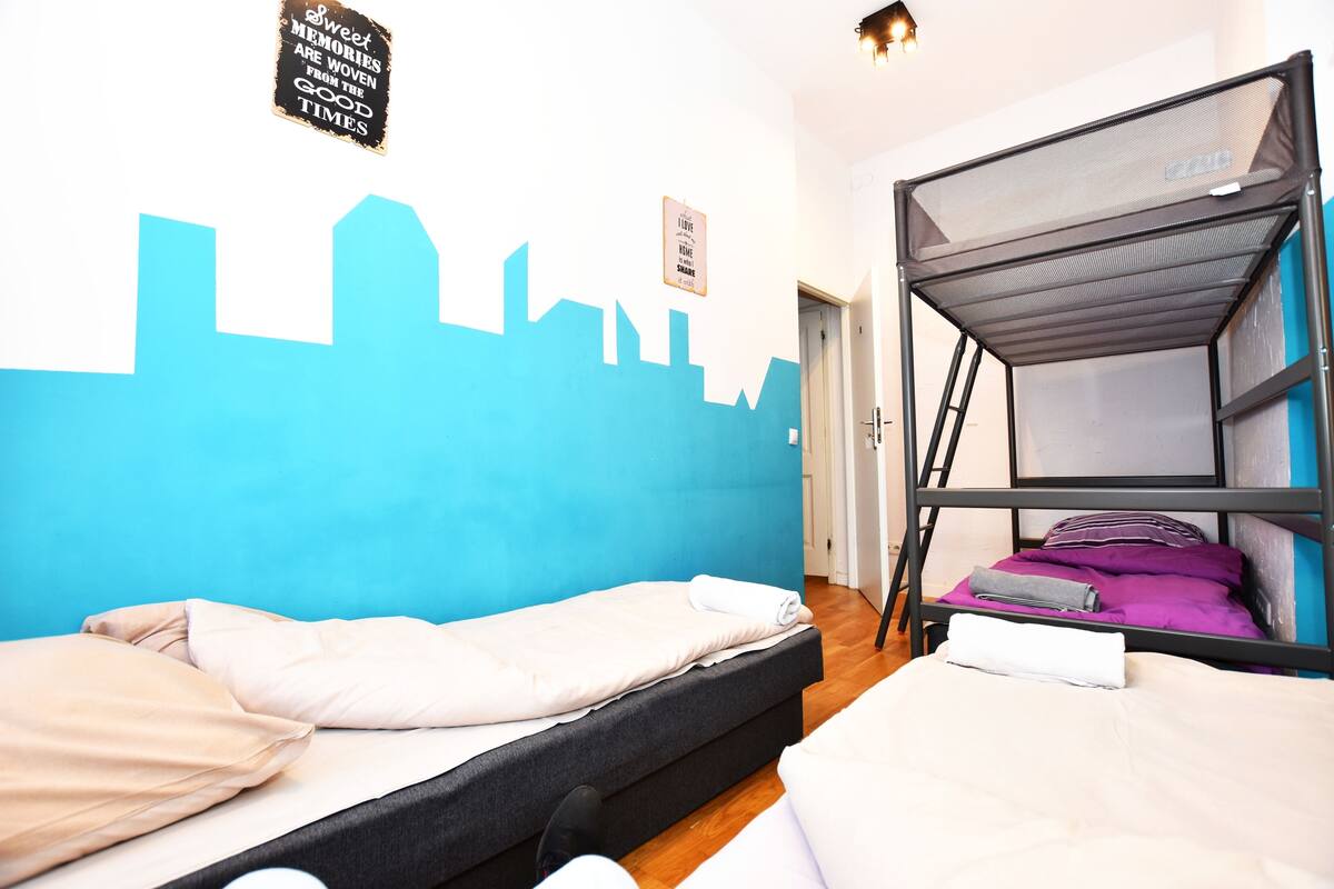 Une chambre avec un lit, un lit superposé et un mur orné d’une fresque représentant un paysage urbain.