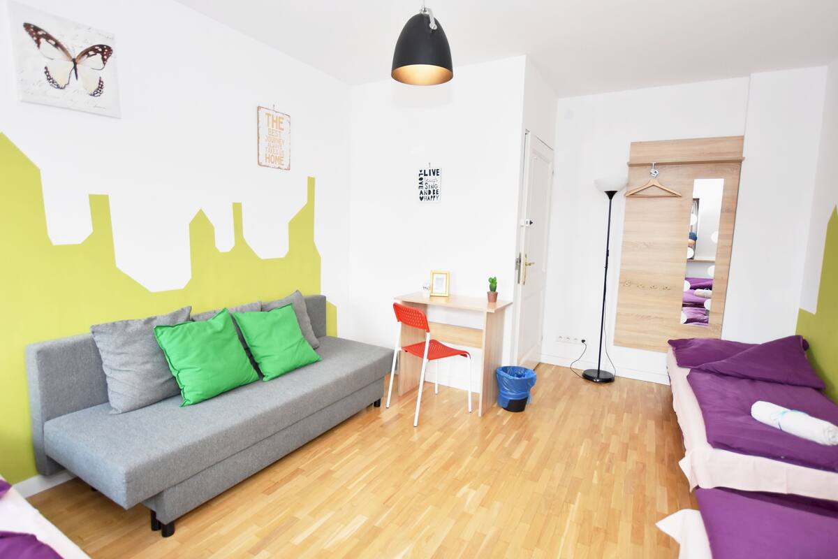 Une chambre moderne avec un canapé gris, un petit bureau, une chaise, un luminaire suspendu et un lit avec une literie violette.