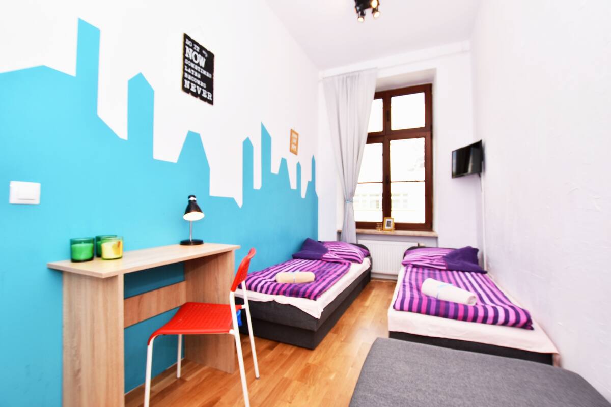 Une chambre avec deux lits, un bureau, une chaise et une fenêtre avec des rideaux.
