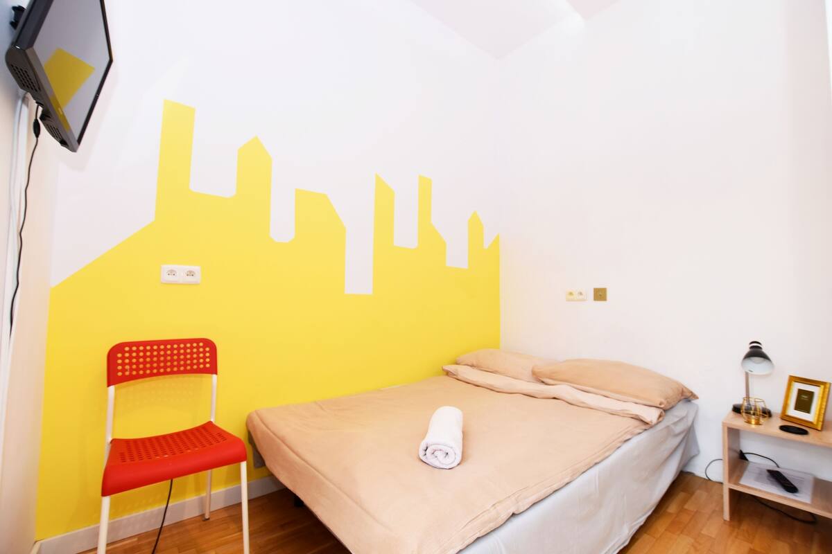 Une pièce comprenant un lit, une chaise, un bureau avec une lampe et un mur décoré d’un motif représentant un paysage urbain jaune.