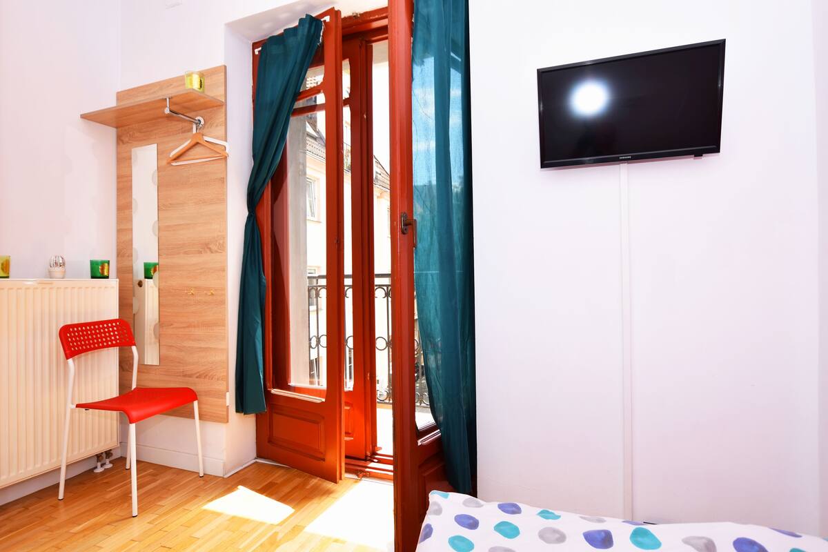 Une pièce avec un plancher en bois, une chaise rouge, un lit recouvert d’un tissu à pois, un téléviseur fixé au mur et une porte ornée de rideaux turquoise.