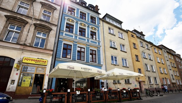 Une rue avec des terrasses de café, une devanture de magasin bien visible avec une enseigne, et un bâtiment dont la façade bleue est facilement reconnaissable.