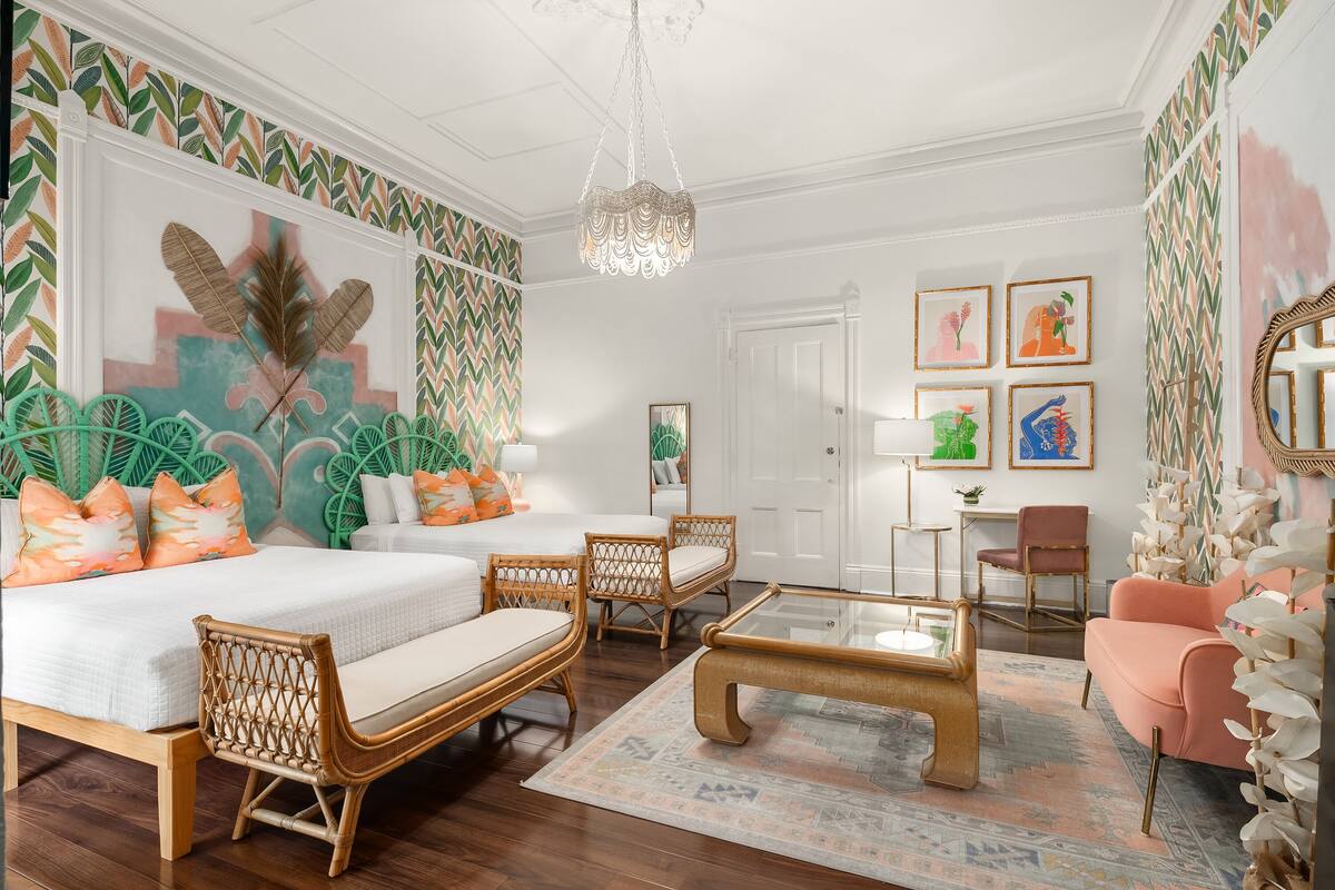 A well-lit room with a bed, two wicker chairs, a coffee table, and a pink armchair. There are framed pictures on the wall and a door in the background.