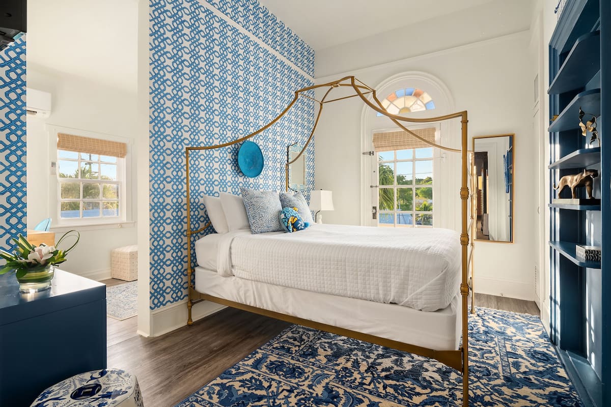 A bedroom with a four-poster bed, patterned wallpaper, and a blue and white rug.