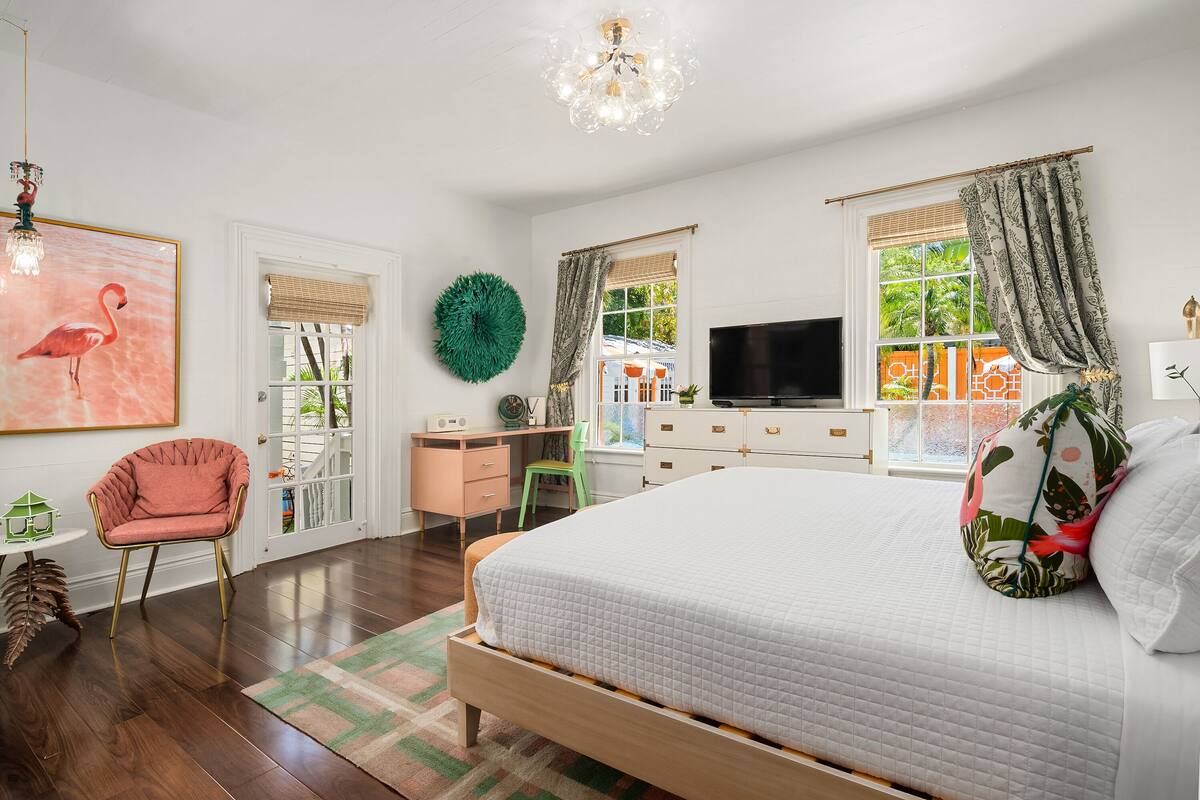 A bedroom with a large bed, a desk, a chair, a television, and a window with curtains.