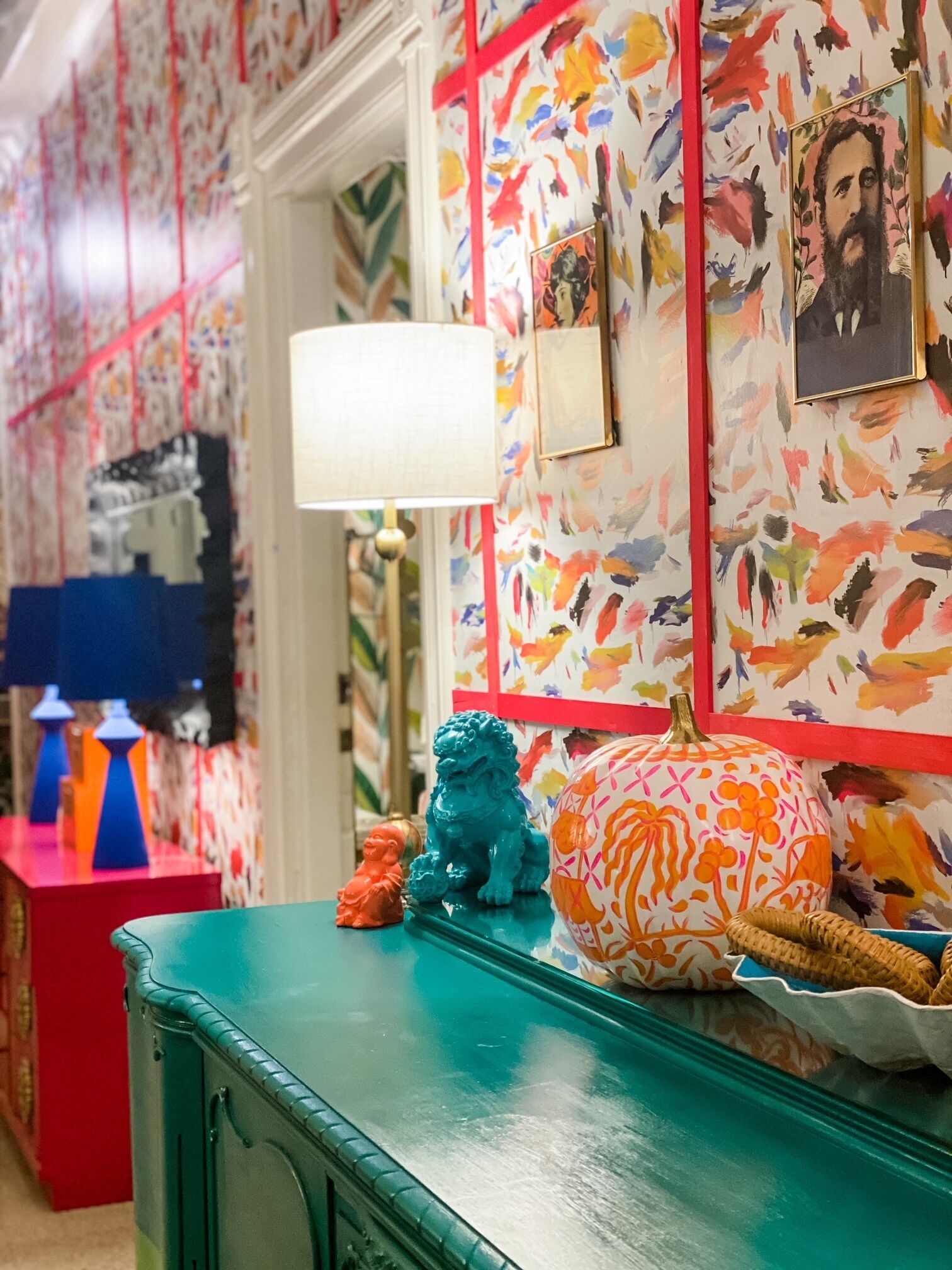 Une pièce avec un papier peint aux motifs floraux éclatants, une commode turquoise et une lampe décorative.