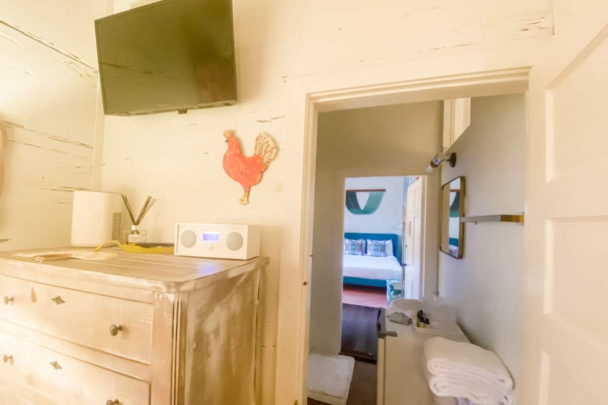 A room with a wooden dresser, a radio, a wall-mounted TV, a rooster decoration, and a doorway leading to another room with a bed and a hanging hammock.