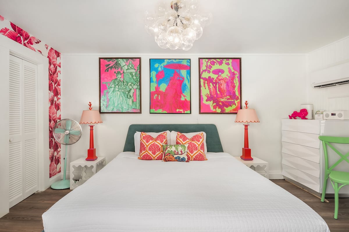 A bedroom with a large bed, two bedside tables, a green chair, and three colorful wall art pieces.