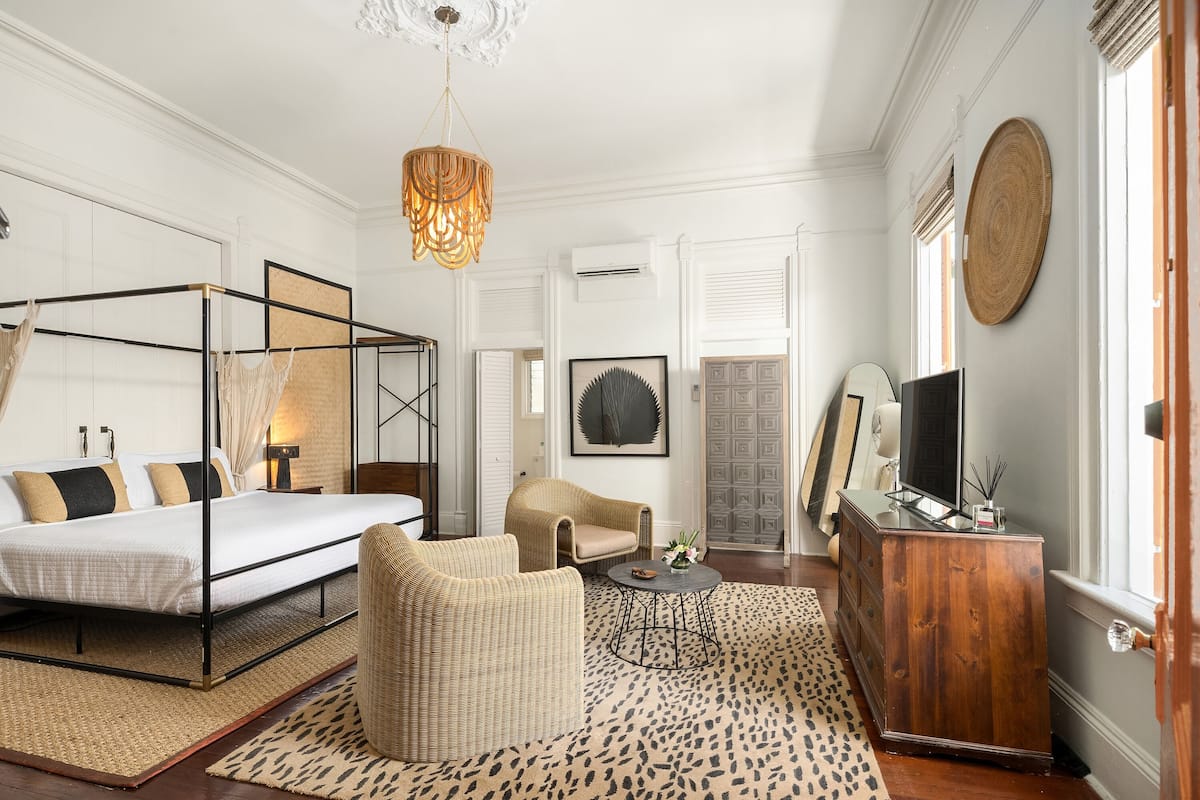A bedroom with a four-poster bed, a wicker chair, a wooden dresser, and a television.