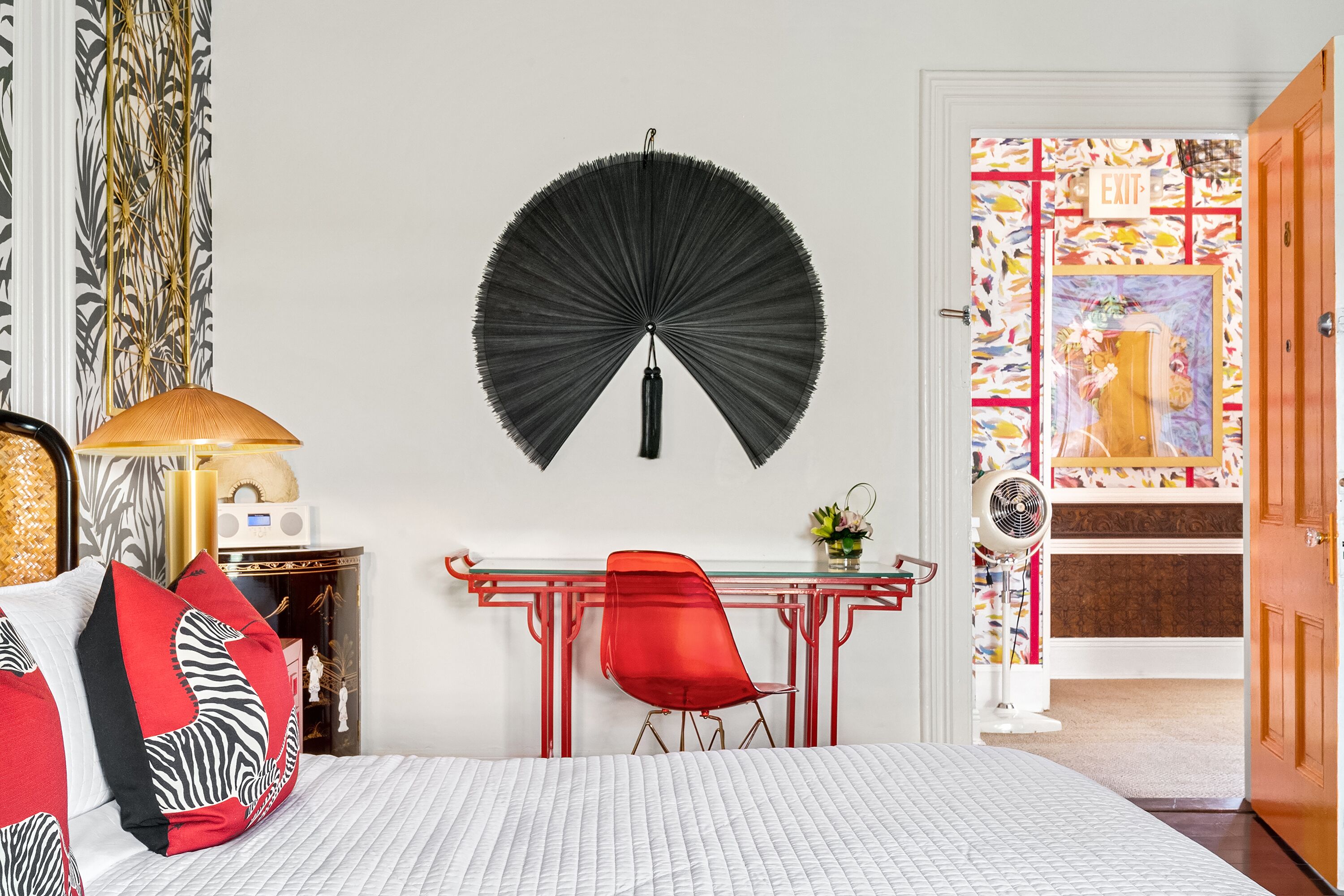 Une chambre à coucher comprenant un lit, un bureau, une chaise, un ventilateur et une porte donnant sur une autre pièce.