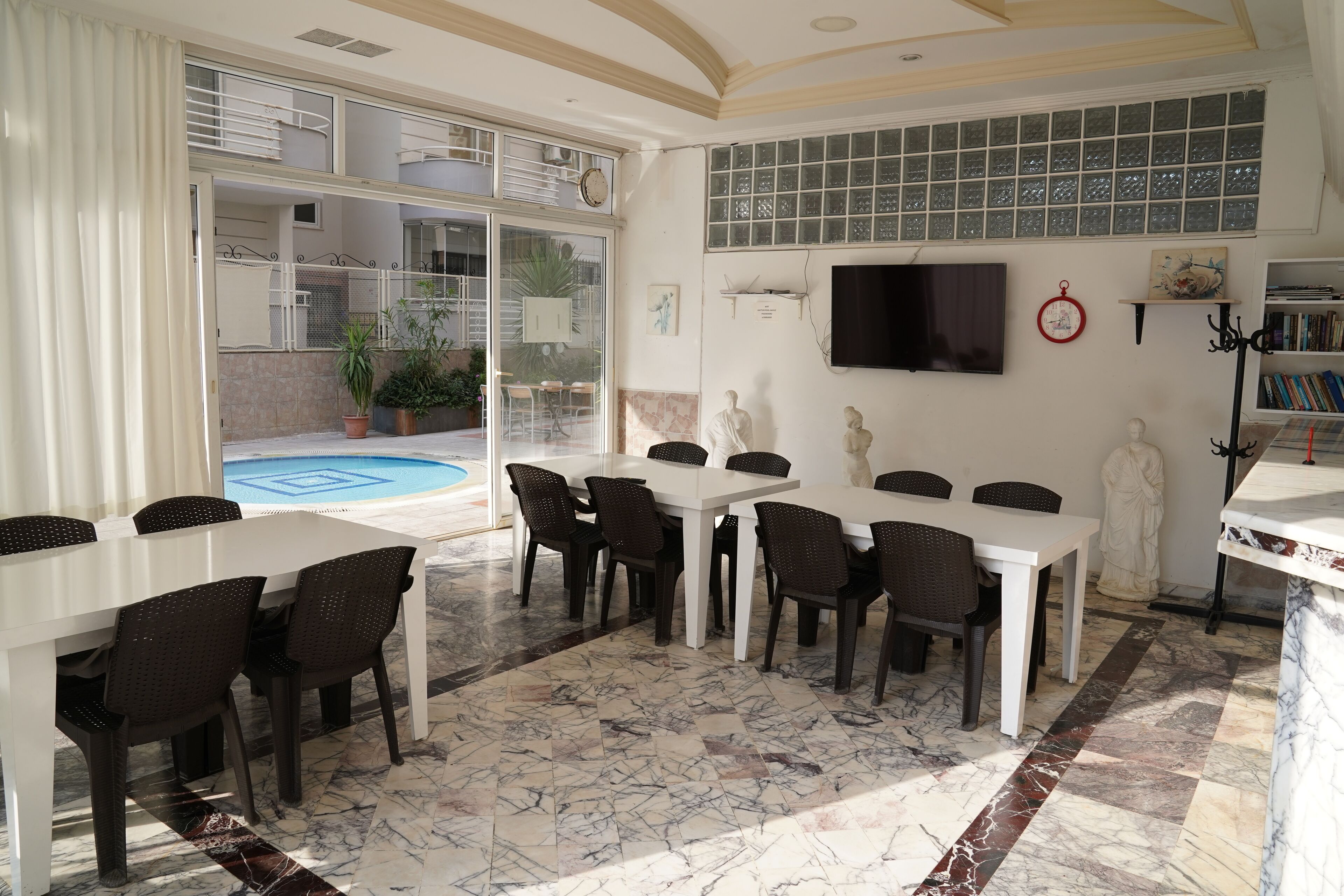 A dining area with white tables and dark chairs, a TV mounted on the wall, and a pool visible through the glass doors.