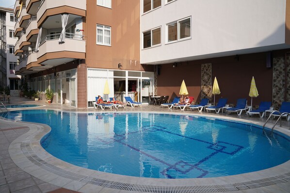 A swimming pool with lounge chairs and umbrellas, surrounded by a building with balconies and windows. - Alanya Polat Tatil Evleri (Alanya)