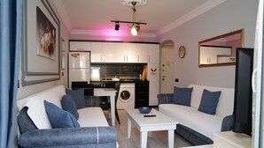 A modern living room with a sofa, a coffee table, and a TV. There is a kitchen in the background with a washing machine and a dining area. - Alanya Polat Tatil Evleri (Alanya)