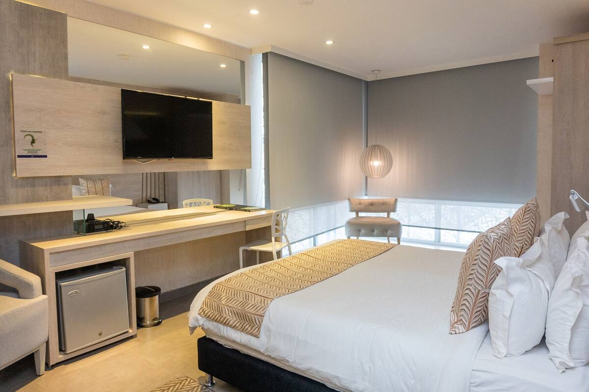 A modern hotel room with a large bed, a desk with a computer, a flat-screen TV, and a small minibar.