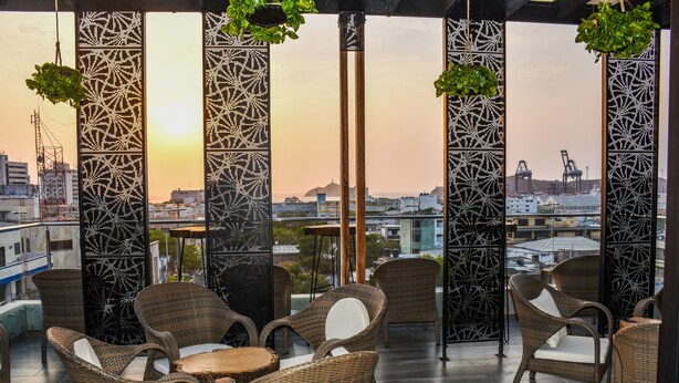 A rooftop terrace with wicker furniture, a patterned column, and a view of the cityscape at sunset. - Hotel Perla Suite Santa Marta (Santa Marta)