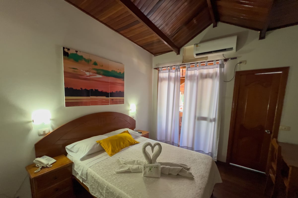 Una habitación de hotel con cama, mesita de noche, un cuadro en la pared, ventana con cortinas y aire acondicionado.
