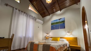 Un dormitorio con cama, mesita de noche, piso de madera, ventilador de techo, ventana con cortinas y un cuadro en la pared.