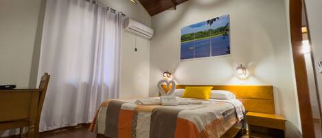 Un dormitorio con cama, mesita de noche, piso de madera, ventilador de techo, ventana con cortinas y un cuadro en la pared.