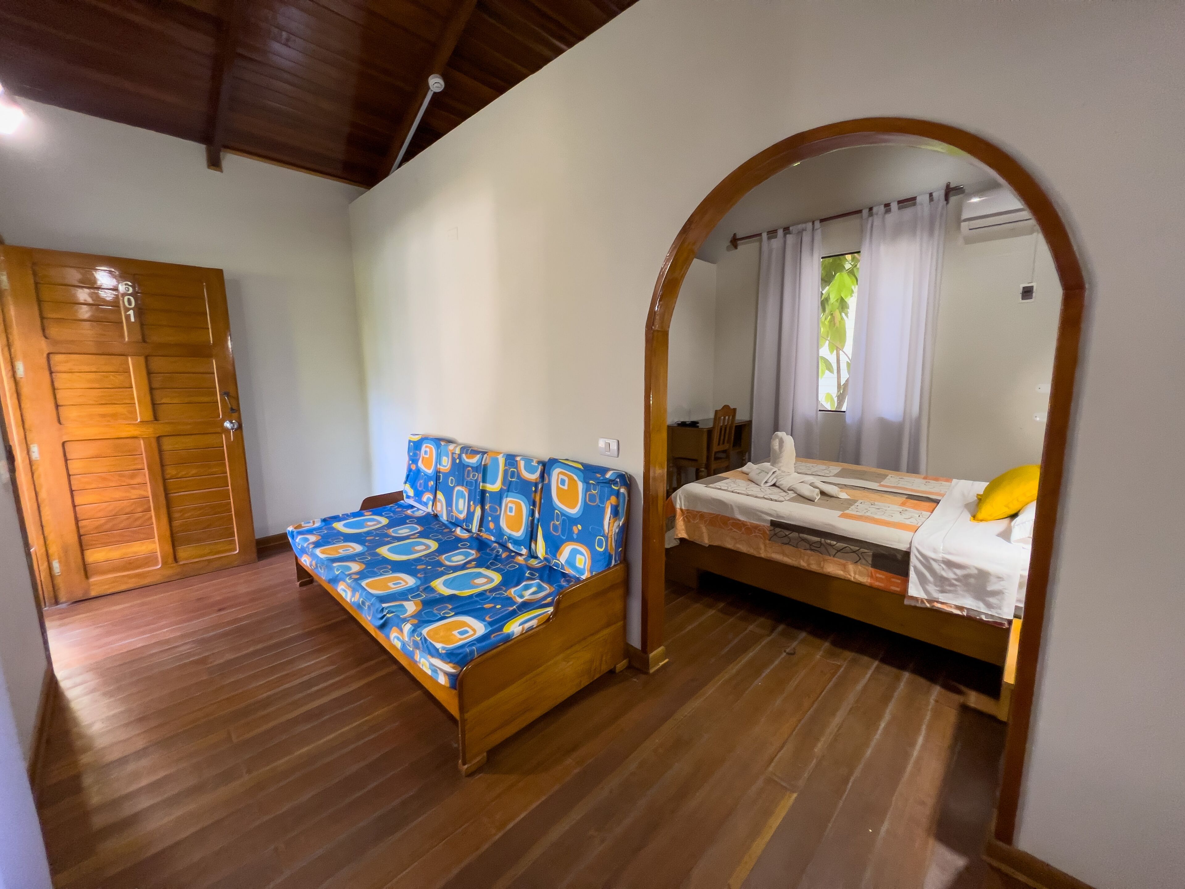 Una habitación con piso de madera, una cama con cubrecama blanco y un sofá azul estampada.