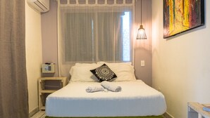 A bedroom with a bed, curtains, an air conditioning unit, a bedside table, a lamp, and a painting on the wall. - Wonderland Hostel By GB Collection (Cartagena)