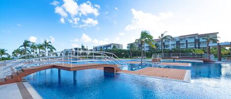 A large outdoor swimming pool with a wooden deck and a bridge, surrounded by palm trees and modern buildings.