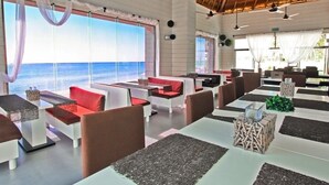 Un restaurante con mesas blancas y sillas rojas y marrones, grandes ventanales con vistas al océano y un techo de paja.