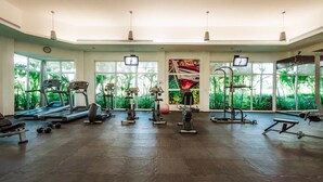 Un gimnasio espacioso con diversos equipos de ejercicio, amplias ventanas con vistas a zonas verdes y un reloj en la pared.