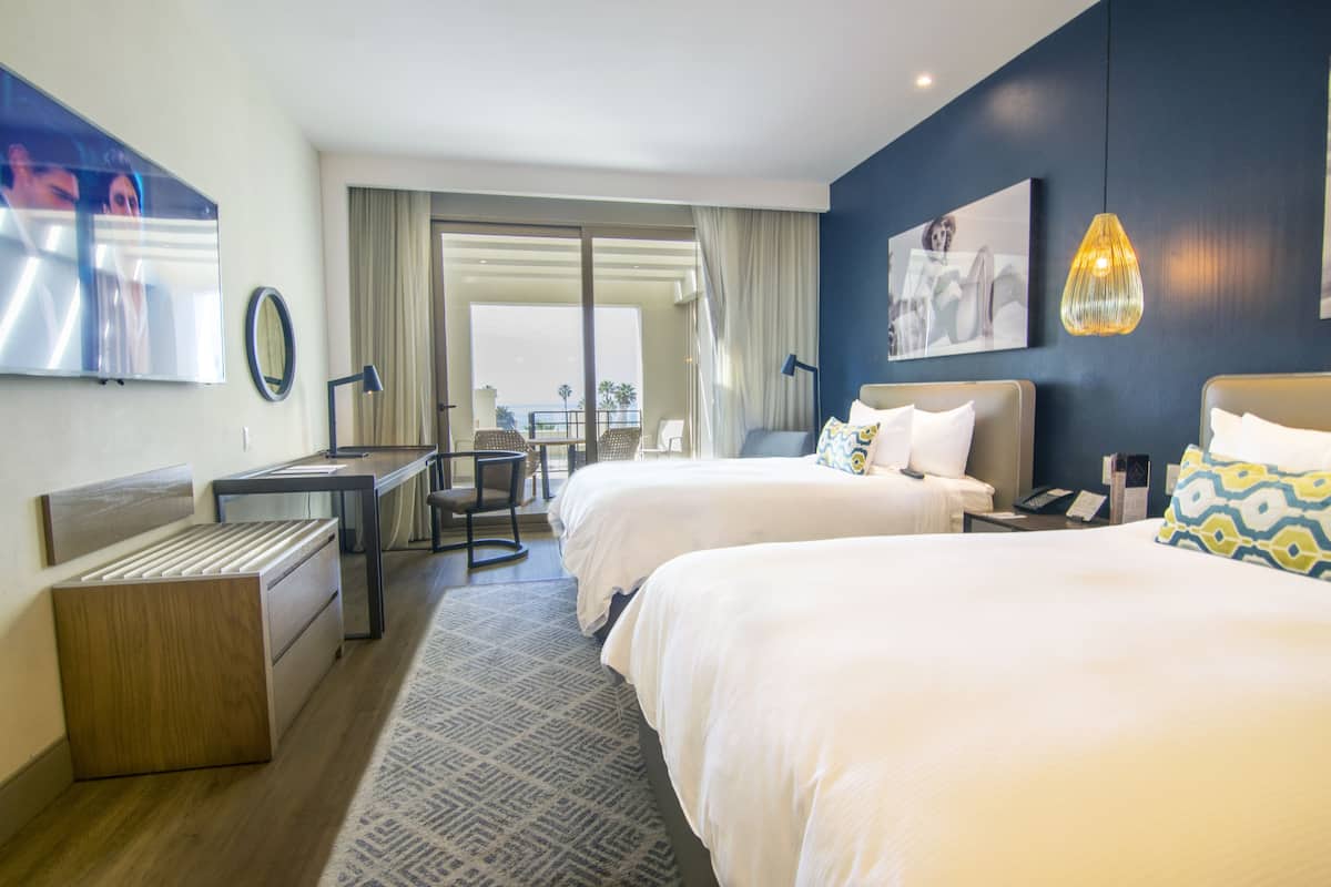 A hotel room with two beds, a desk, a chair, a TV, and a view of the outdoors.
