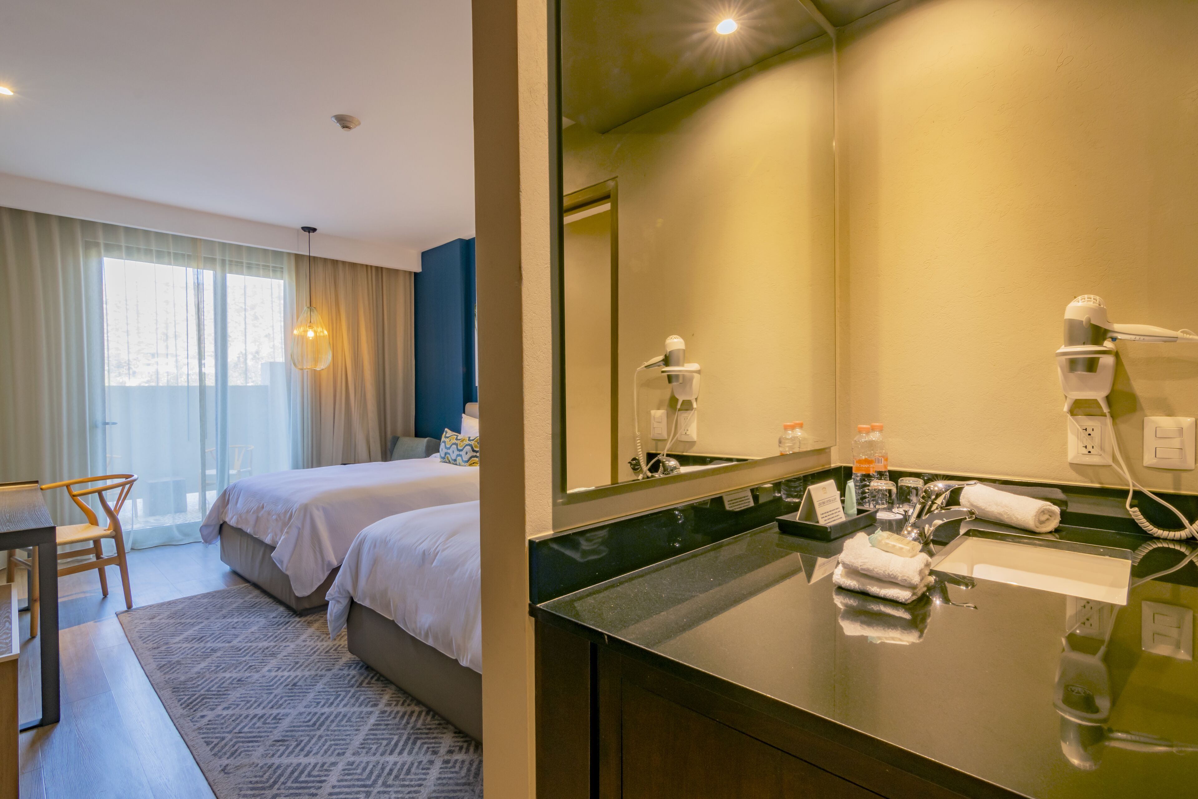 A hotel room with a large bed, a desk, a chair, a bathroom with a mirror, a sink, and a towel rack.