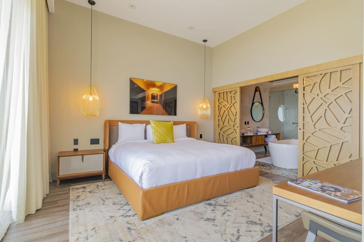 A modern hotel room with a large bed, bedside tables, a bathroom with a bathtub, and a wooden partition.